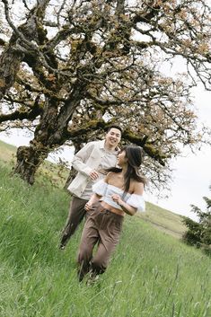 a man and woman are walking through the grass in front of a tree with no leaves