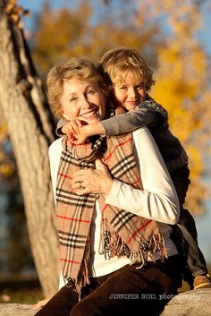 an older woman and young boy sitting on the ground in front of a tree with autumn leaves