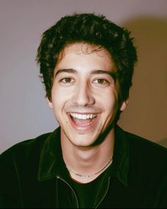 a man with curly hair smiling at the camera while wearing a black shirt and silver necklace