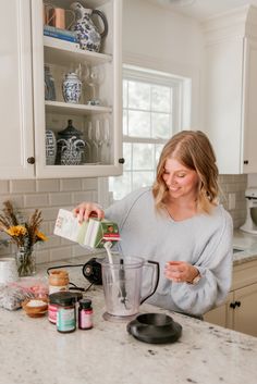 a woman pouring something into a blender in a kitchen with white cabinets and marble counter tops