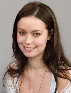 a woman with long brown hair wearing a white shirt and necklace smiling at the camera