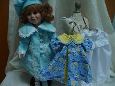 two dolls are sitting next to each other on a white cloth covered surface, one is wearing a blue coat and the other has a yellow dress