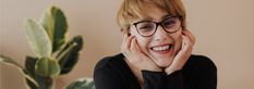 a woman with glasses smiling at the camera while leaning on her hand near a potted plant