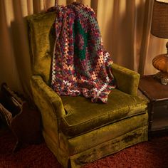 a green chair with a crocheted blanket on it next to a table and lamp