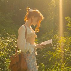 a woman standing in the grass holding a book and looking at it with sunlight shining on her face