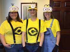 three women in yellow shirts and overalls wearing minion hats with googly eyes