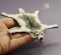 a hand holding a tiny toy animal on top of a purple surface with white and gray fur