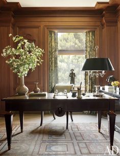 a desk with a lamp, vase and flowers on it in front of a window