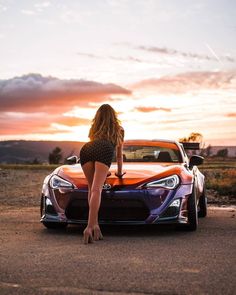 a woman leaning on the hood of a sports car in front of a beautiful sunset