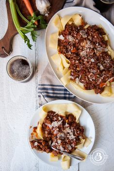 two bowls filled with pasta and meat on top of a white table next to vegetables