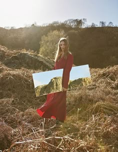 a woman in a red dress is holding a mirror and posing for the camera while standing on a hill