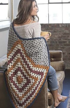 a woman sitting on a couch holding a coffee cup and wearing a crocheted blanket