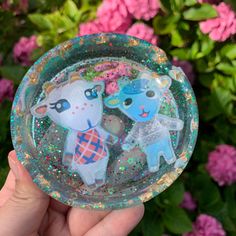 someone holding up a clear glass bowl with two small animals in it and pink flowers behind them