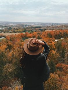 a woman wearing a hat looking out over the trees in an open field with autumn foliage