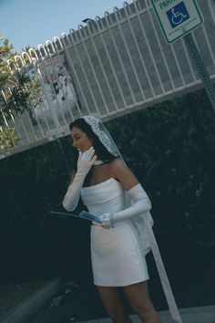 a woman dressed in white is standing on the sidewalk with her veil over her face