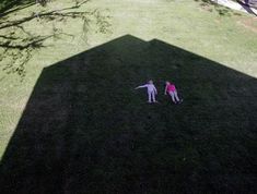 two people are standing in the shadow of a large object on grass, with trees behind them