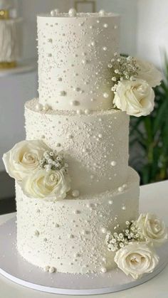 a three tiered wedding cake with white roses and pearls on the top, sitting on a table