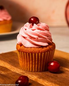 a cupcake with pink frosting and cherries on a cutting board next to a plate