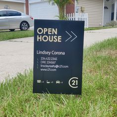 an open house sign sitting in the grass next to a driveway with a car parked behind it