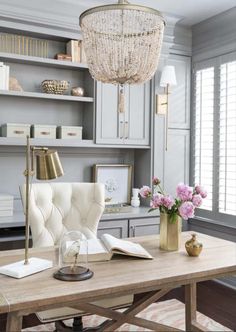 a desk with a chair, lamp and flowers on it in front of a bookshelf