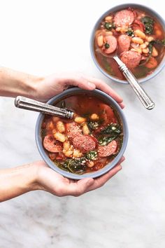 two hands holding bowls of sausage and bean soup