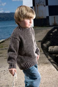 a young boy walking on the beach with a frisbee in his hand while wearing a sweater and jeans