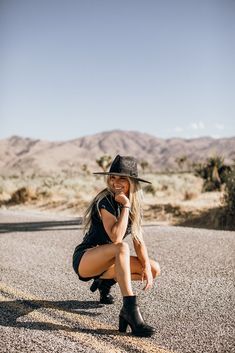 a woman sitting on the side of a road wearing a cowboy hat and black dress