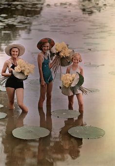 three women in swimsuits and hats standing on the beach with water lillies