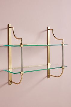 two glass shelves on the wall with brass brackets and hooks attached to them, against a pink wall