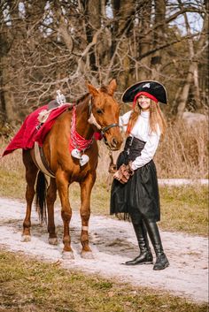 a woman in pirate garb standing next to a brown horse with a red blanket on it's back