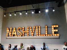 people sitting on benches in front of a sign that reads nashvillee with lights hanging from the ceiling