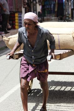 an old man carrying wood on his back down the street with a cart behind him