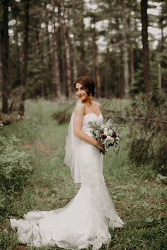 a woman in a wedding dress is standing in the woods holding a bouquet and looking at the camera