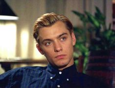 a man with blonde hair and blue eyes sitting in a chair next to a potted plant