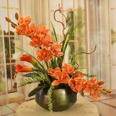 an arrangement of orange flowers in a green vase on a table with windows behind it