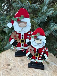 two santa claus figurines sitting on top of a wooden table next to a pine tree