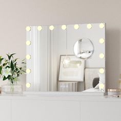 a white dresser with a mirror and lights on it in front of a vase filled with flowers