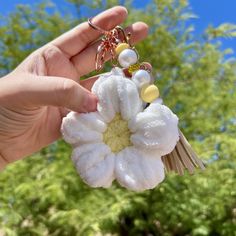 a hand holding a white flower keychain in front of some trees and bushes