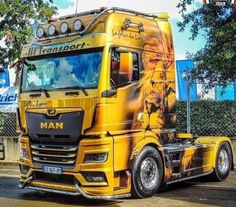 a yellow semi truck parked in front of a fenced area with trees and bushes