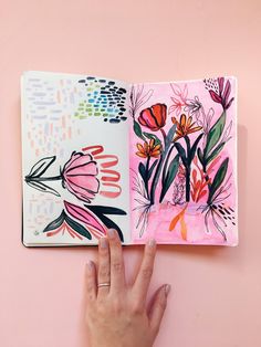 a person's hand is holding an open notebook with flowers on it and pink background