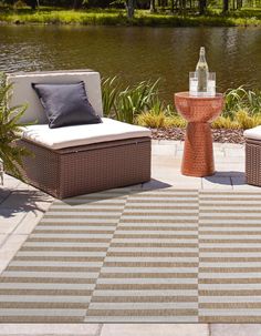 two wicker lounge chairs sitting on top of a patio next to a lake with a bottle of wine