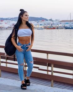 a woman in ripped jeans standing on a pier next to the water with her back turned