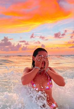 a woman covers her eyes as she stands in the ocean at sunset with an orange and pink sky behind her