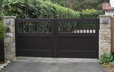 a driveway with a black gate and stone pillars in front of a house surrounded by greenery