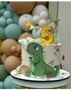 a birthday cake with a dinosaur on top and the number five in the middle, surrounded by balloons