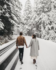 a man and woman holding hands while walking down a snow covered road in the woods