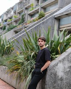 a man leaning against a stone wall with plants growing on the side and buildings in the background