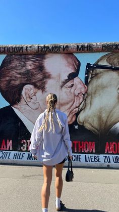 a woman standing in front of a wall with a painting of two men kissing each other