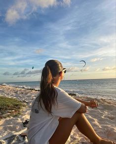 a woman sitting on top of a sandy beach next to the ocean with kites flying in the sky