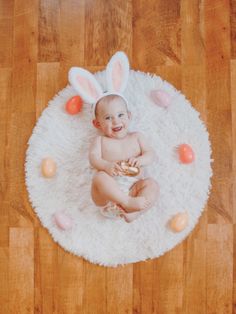 a baby in bunny ears is sitting on a rug with eggs around him and smiling at the camera